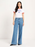 A model stands against a plain light background wearing a white ringer T-shirt with red trim and blue collegiate-style text across the chest, paired with high-waisted light blue wide-leg jeans.