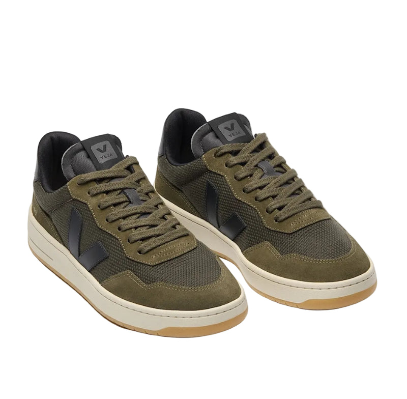 A pair of low-top sneakers in olive green and dark gray, featuring a textured mesh and suede upper, matching green laces, and a cream midsole with a gum outsole, shown on a light gray background.
