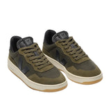 A pair of low-top sneakers in olive green and dark gray, featuring a textured mesh and suede upper, matching green laces, and a cream midsole with a gum outsole, shown on a light gray background.